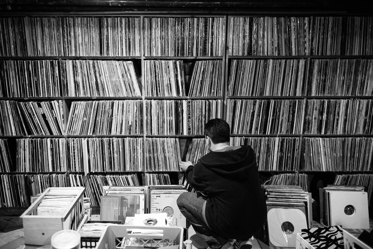 Black and white photo of a wall of shelves filled with records. Someone is crouched on the ground pulling a record out, crates of records are on the floor.