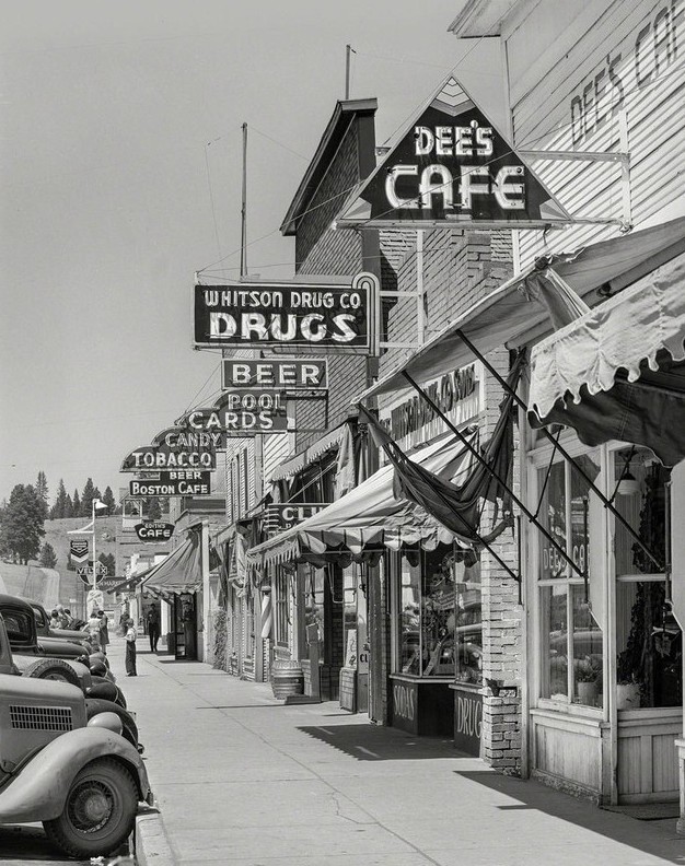 A vintage photograph looking down a small-town sidewalk lined with storefronts and stacked neon signs, including Dee's Cafe, Whitson Drug Co., Beer, Cards, Candy, Tobacco, and Boston Cafe.