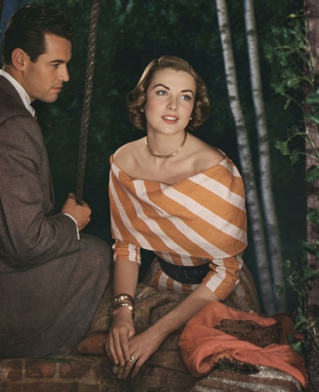 A woman with styled short brown hair sits outdoors wearing an off-the-shoulder orange and white striped top and a gold choker necklace. Beside her, a man in a grey suit looks toward her.