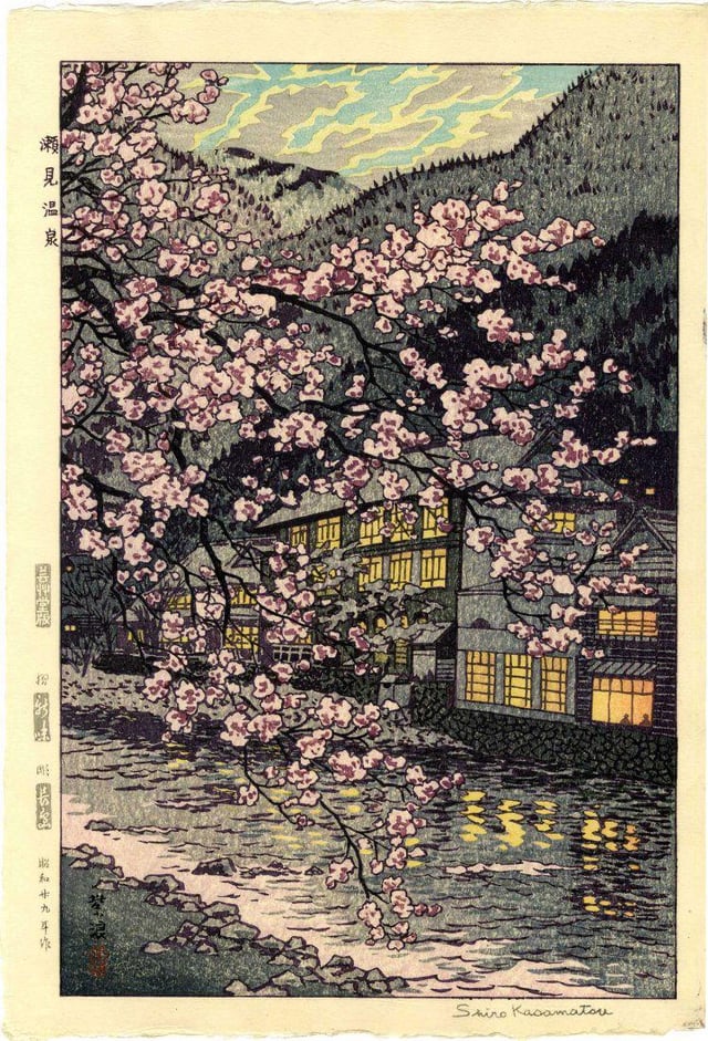 Pink cherry blossoms dominate this woodcut frame, behind a building at the foot of a mountain, yellow light from its windows