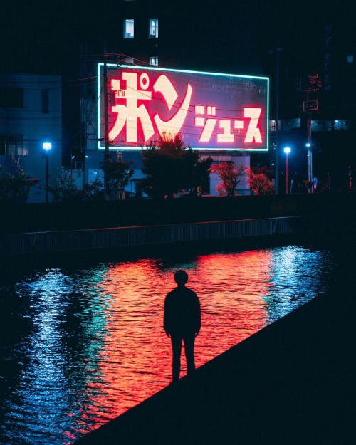 A lone silhouette figure stands in front of a river that is reflecting a lighted street sign with Japanese lettering that is casting a red light