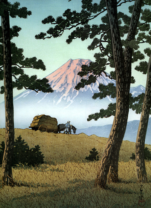 A woodblock scene with Mt. Fuji in the background. A figure leads a horse and hay cart in the middle distance. A few trees and grassy field are in the foreground.