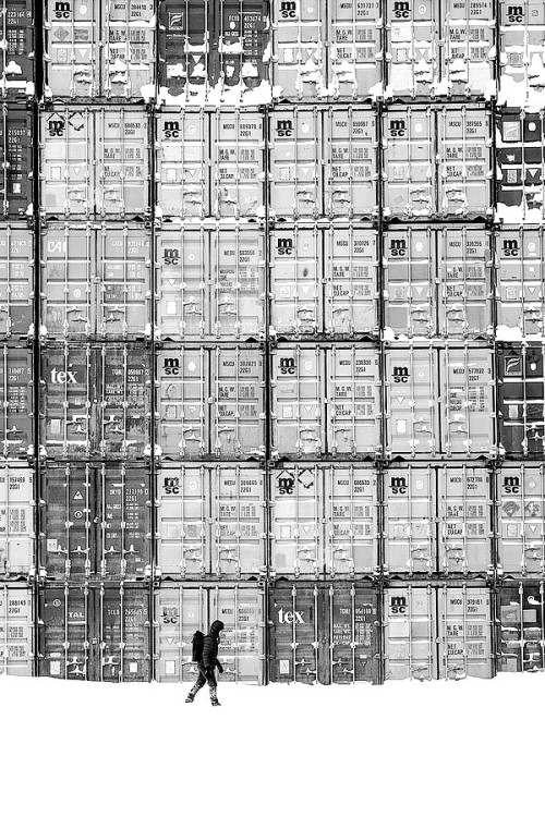 A snowy scene with stacks of shipping containers. A small, bundled lone figure with a backpack is walking in front and shows the scale.