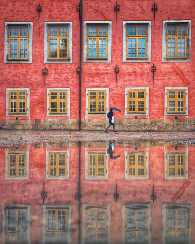 A photograph of a pink building with yellow-paned windows reflecting in water, a lone figure walks in front with an umbrella 