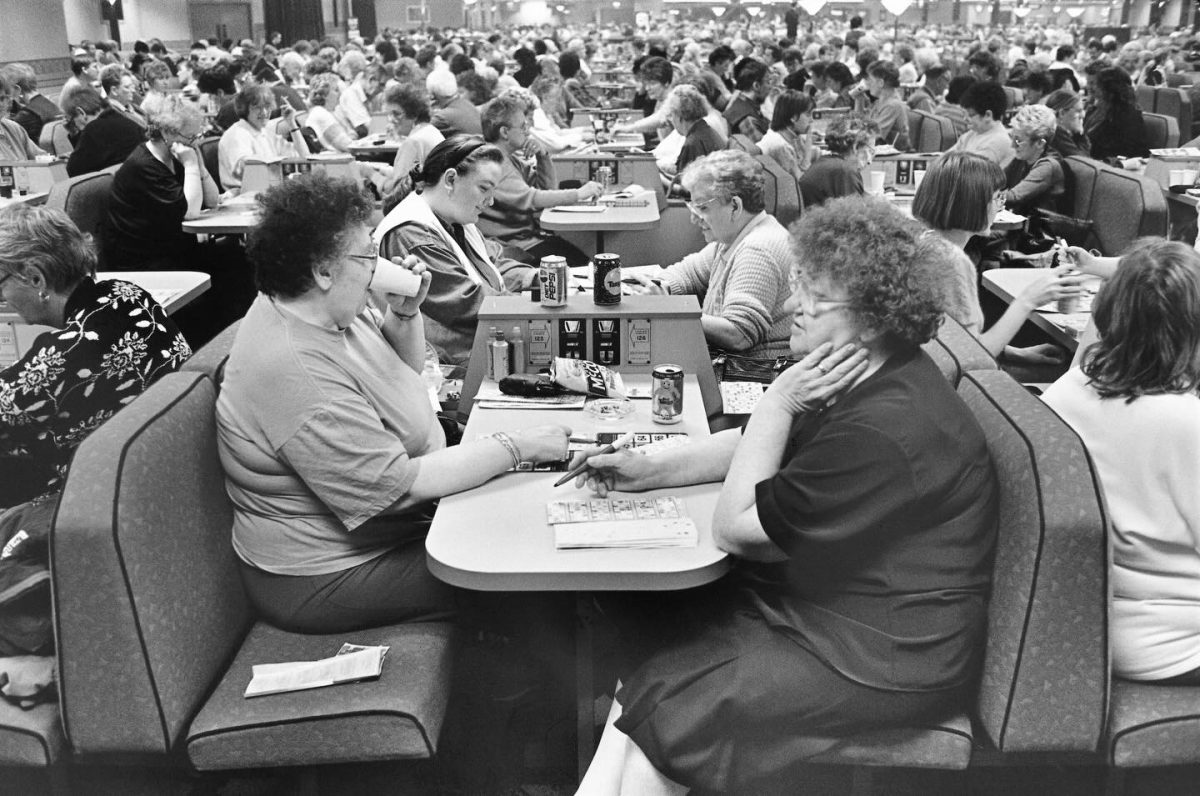 A black-and-white, eye-level photograph of a crowded bingo hall, capturing numerous people sitting in booths at tables, focused on playing the game.
