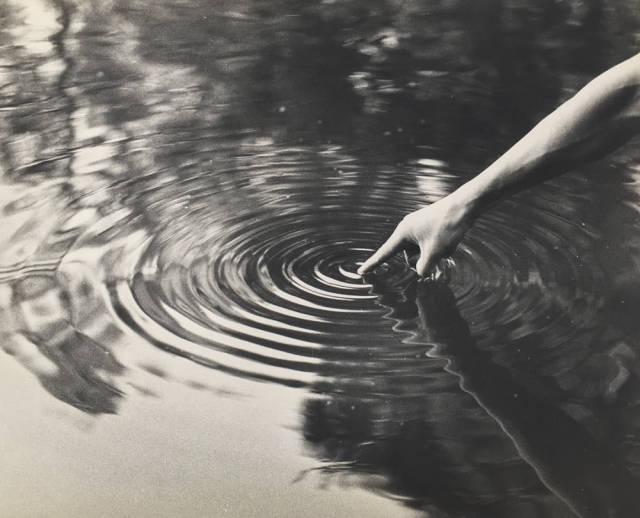 Black and white photo of a finger touching water creating ripples, with trees reflecting