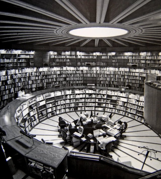 A black-and-white, high-angle photograph of a circular library featuring multiple tiers of curved bookshelves and a central workspace with people at computers.