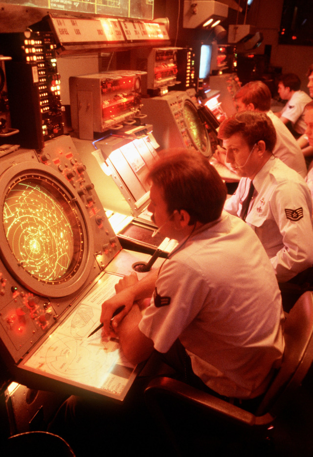 Military personnel sit at complex electronic monitoring consoles in a dimly lit room filled with amber and red light. In the foreground, an operator views a circular radar screen filled with data blips while working on a lit display table.