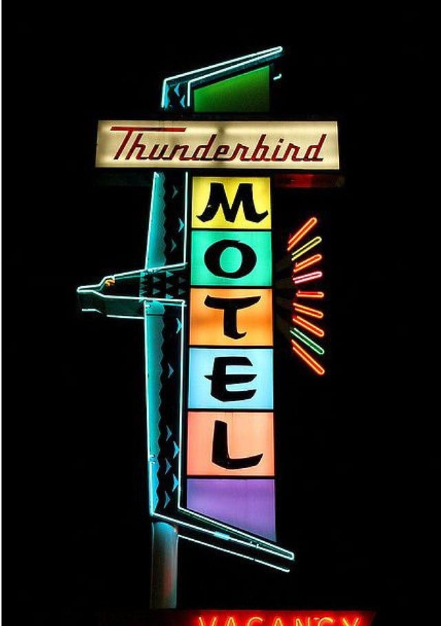 A vintage neon sign for the Thunderbird Motel at night. The word "Thunderbird" in red script sits above a vertical stack of colored blocks spelling "MOTEL". To the left, a stylized neon outline forms a thunderbird.