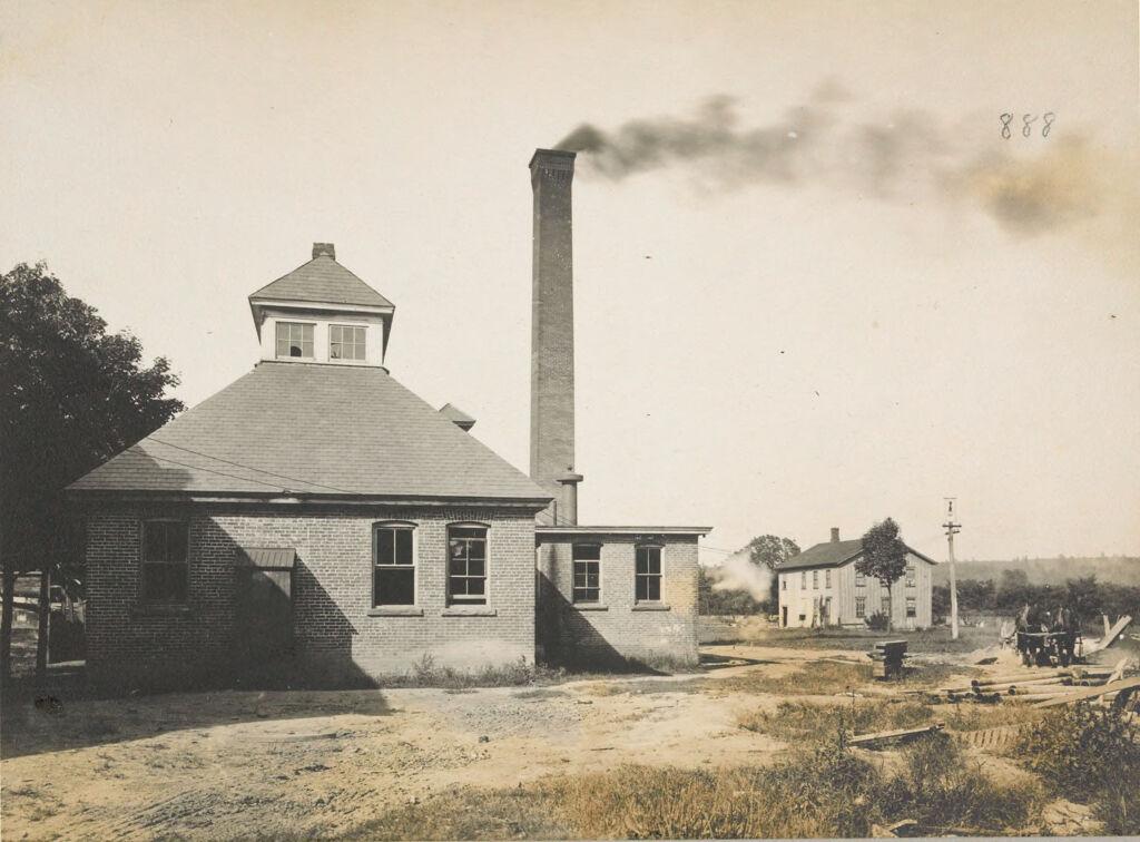 A sepia-toned photograph of a brick industrial building with a tall smokestack emitting dark smoke, featuring a horse-drawn wagon and another building in the rural background