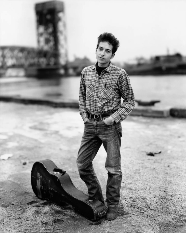 Black and white photo of a young Bob Dylan in a plaid shirt and jeans stands with his hands in his pockets and a guitar case at his feet on an industrial waterfront.
