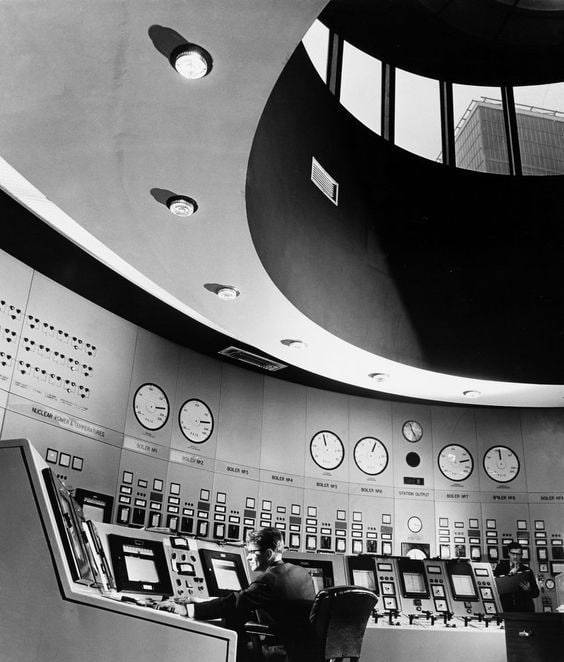 A man sits at a curved control console in a mid-century industrial facility. The room features expansive banks of analog dials and a dramatic circular ceiling opening that looks out onto another building.