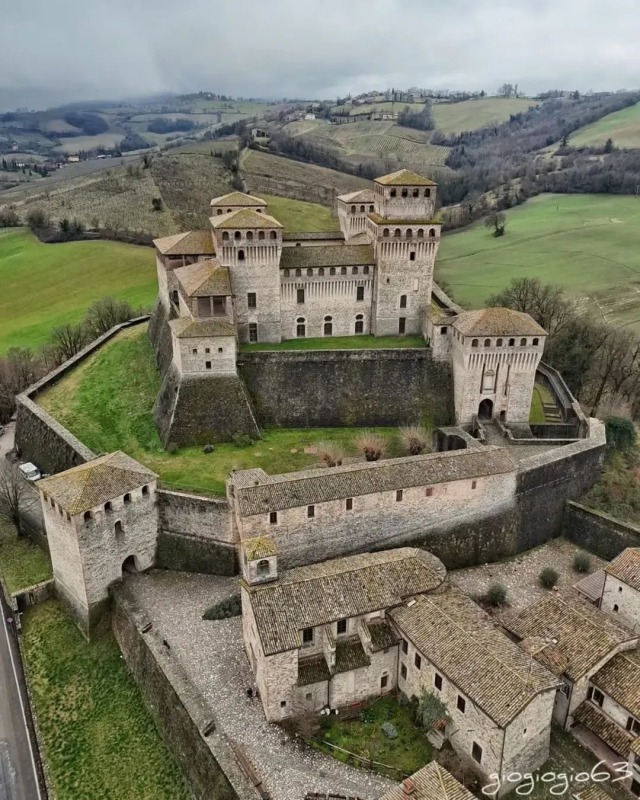 Aerial view of a large stone medieval castle with multiple towers and clay tile roofs, situated on a grassy hill overlooking a rolling green landscape under an overcast sky.