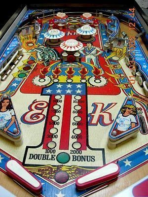 A vintage Evel Knievel pinball machine playfield with a red, white, and blue patriotic theme. The center features a large number 1 decorated with stars and stripes flanked by the letters E and K, surrounded by bumpers, flippers, and illustrations of a stunt motorcyclist.