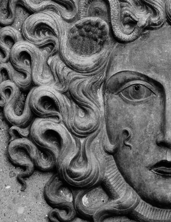 Black and white stone relief sculpture of Medusa, showing half of her face with a wide-open eye and hair made of coiled, writhing snakes.