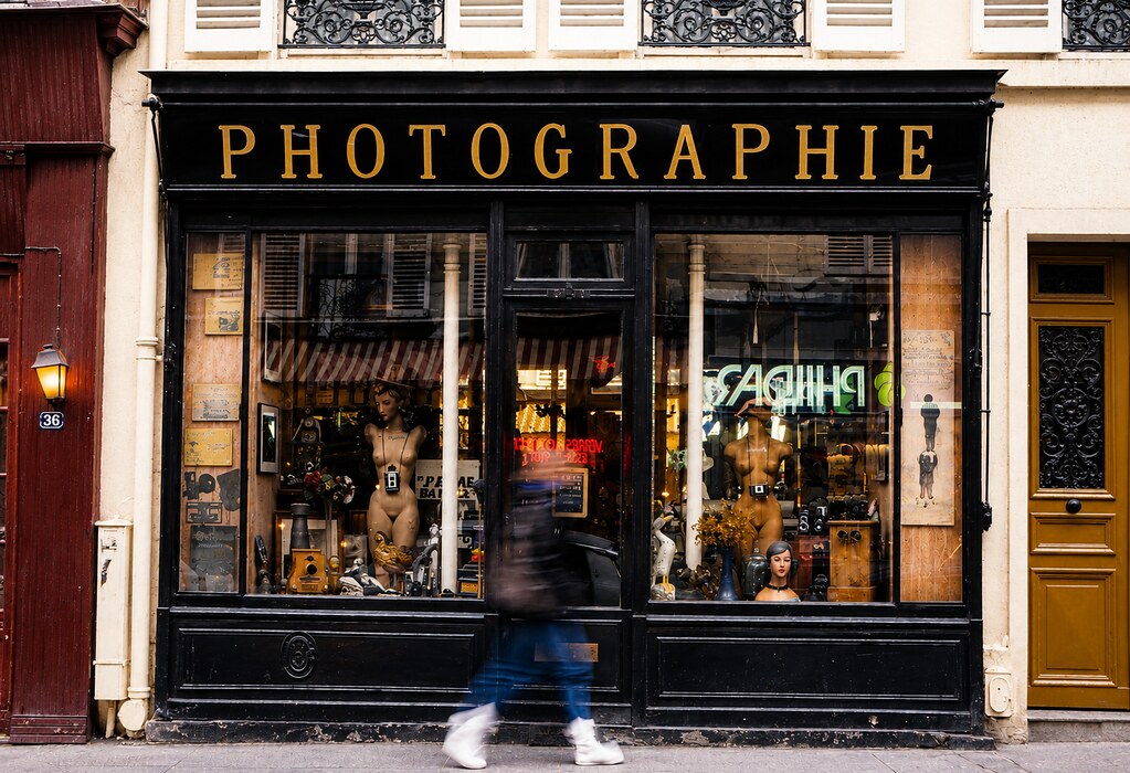 A vintage storefront with a black facade and a gold sign reading "PHOTOGRAPHIE." The large display windows are filled with antique cameras and mannequins. A blurred pedestrian walks past on the sidewalk in the foreground.