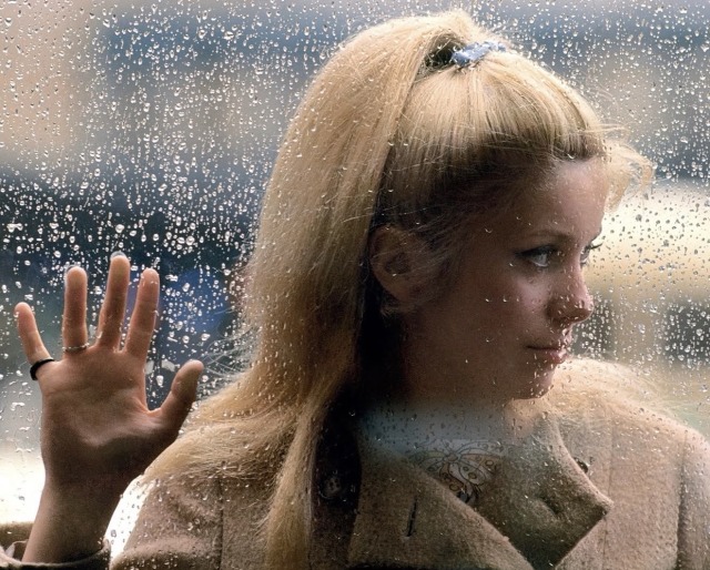 Catherine Deneuve in a beige coat with a 1960s hairstyle looks across a rain-splattered window, pressing her hand flat against the wet glass.
