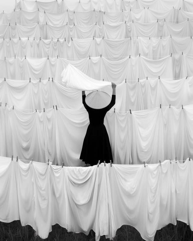 A person in a black dress and a conical hat stands facing away, lifting a white cloth. They are surrounded by tiered rows of hanging white sheets that stretch across the entire frame in a high-contrast black-and-white composition.