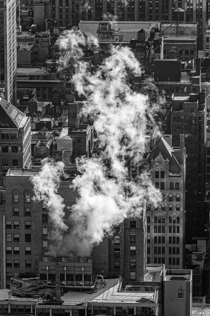 A black and white high-angle view of a dense city, featuring a large, bright plume of white steam billowing upward from a rooftop cooling system in the foreground.