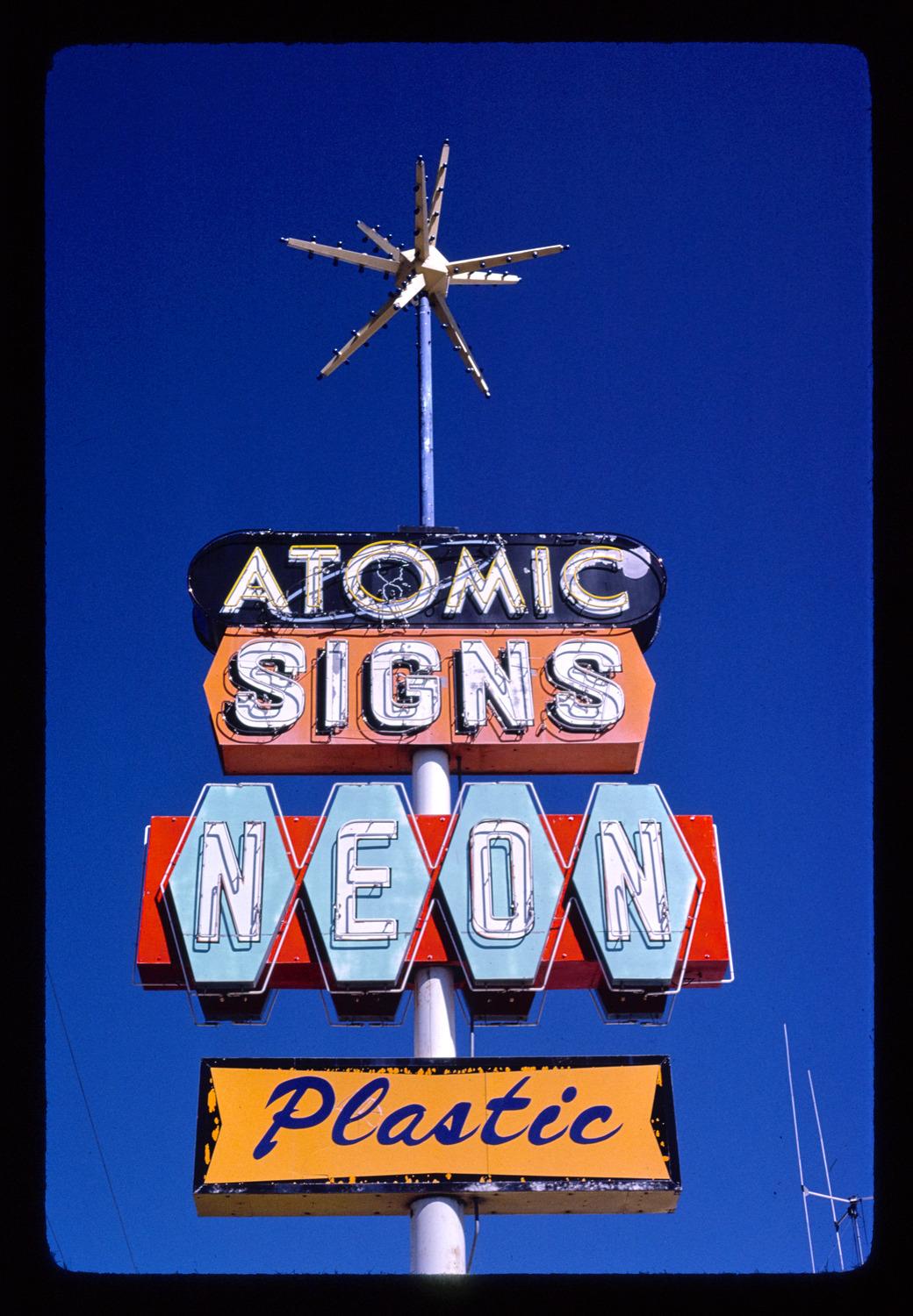 Vintage mid-century modern neon sign against a clear blue sky, topped with a metallic starburst ornament. From top to bottom, the stacked panels read: "ATOMIC" in white neon on black, "SIGNS" in white neon on orange, "NEON" in white letters on light blue shields over a red background, and "Plastic" in blue cursive on a yellow panel.