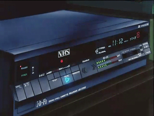 Anime illustration of a vintage dark blue VHS VCR player with a glowing blue play button, digital display reading 11:12, and illuminated audio level meters.