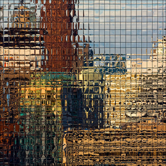 Distorted reflection of city buildings and sky in a grid of square glass window panes, creating an abstract mosaic of gold, brown, and blue.