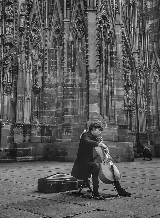 A man plays a cello on a stone-paved plaza in front of the towering, ornate facade of a Gothic cathedral.