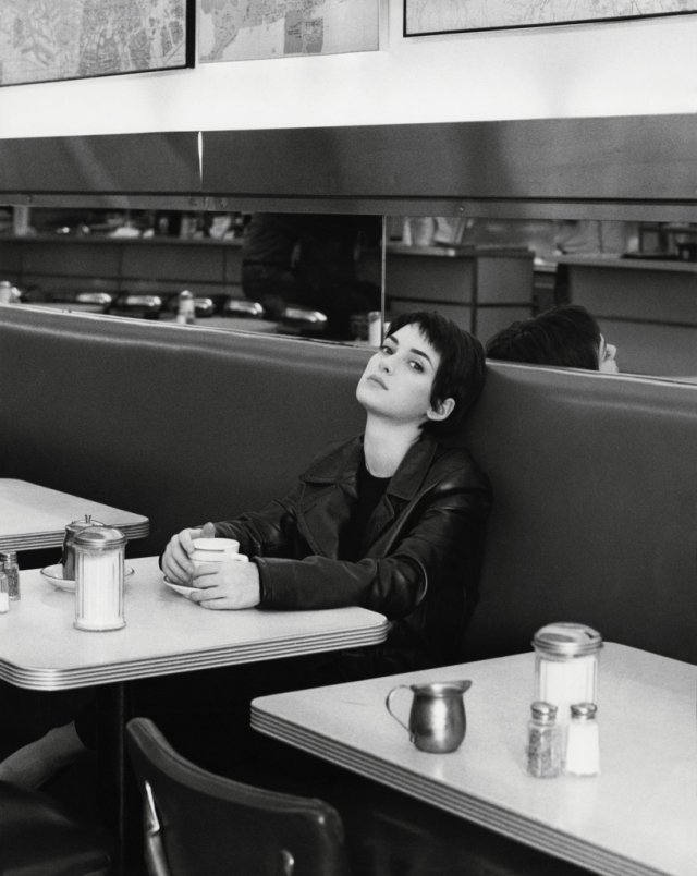 A black-and-white photograph of Winona Rider with short dark hair sitting in a diner booth. She wears a leather jacket, holds a coffee cup with both hands on the table, and leans her head back against the dark cushion, looking directly at the camera.