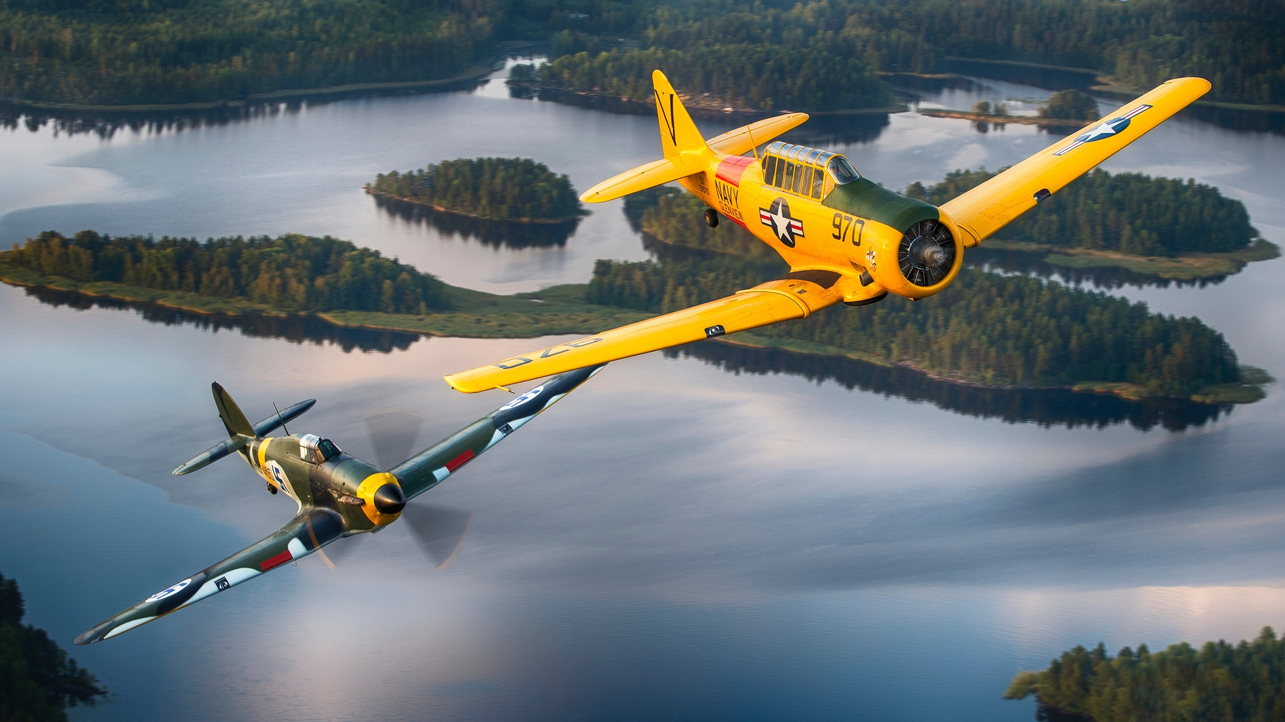 Two vintage military airplanes fly in formation high above a tranquil lake dotted with small, forested islands. A bright yellow US Navy plane banks sharply in the upper right, while an olive-green camouflage fighter plane flies below it to the left.