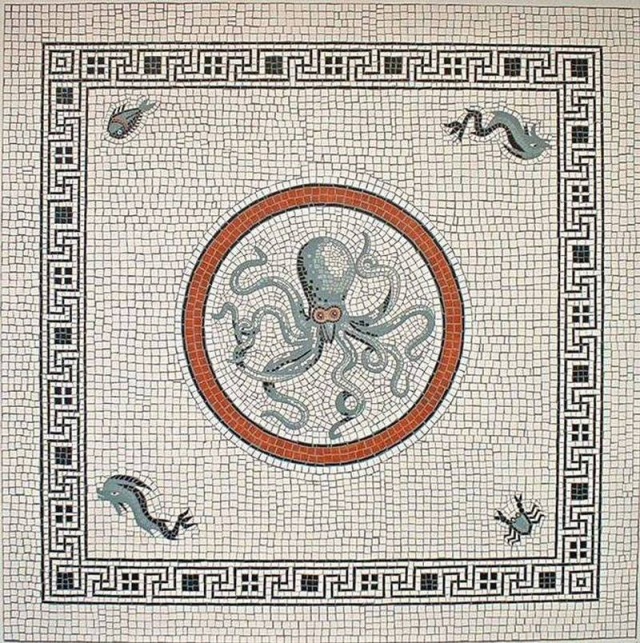A square Roman-style mosaic features a central circular medallion with a terracotta-red border framing a blue-grey octopus with swirling tentacles and wide eyes. Light-colored tesserae fill the background, adorned in each corner with a different sea creature: a small fish in the upper-left, an eel-like sea creature in the upper-right, a dolphin in the lower-left, and a crab in the lower-right. A broad geometric black-and-white Greek key border frames the entire design.