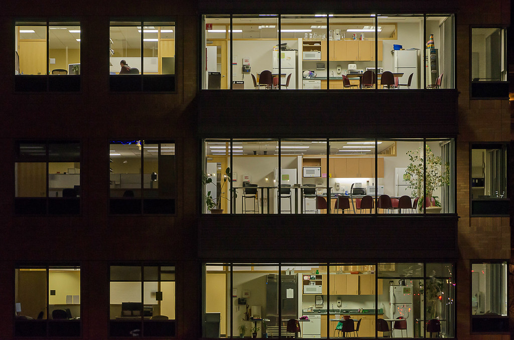 Exterior night view of an office building with illuminated windows across three floors. Larger multi-pane windows reveal nearly identical office breakrooms stacked vertically, each equipped with kitchen cabinets, refrigerators, and dining chairs, while smaller adjacent windows show office cubicles with one person working late.