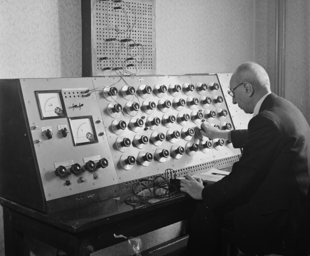 A man wearing a suit and glasses adjusts a knob on an electric panel with around 40 knobs
