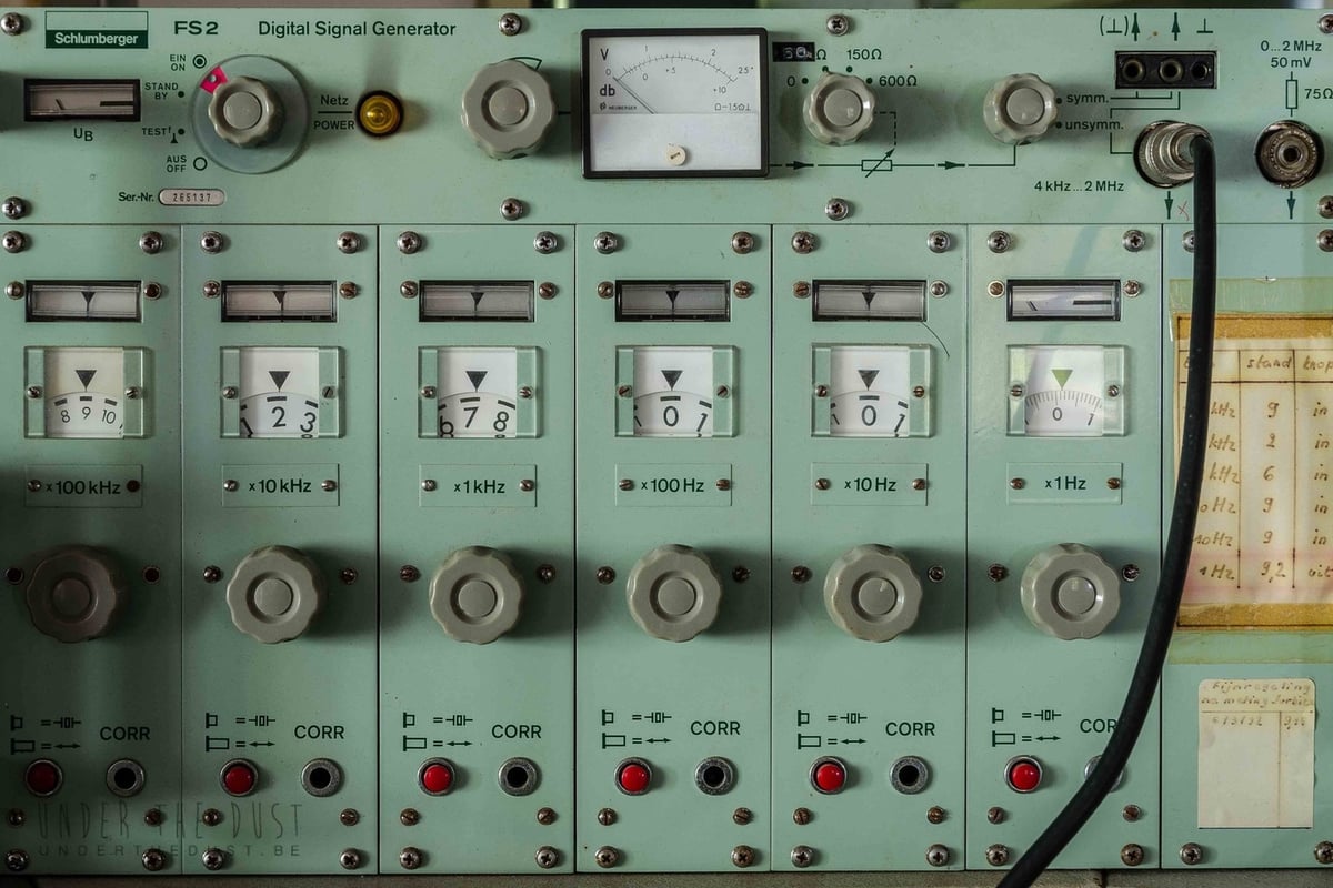 A vintage digital signal generator with a pale green front panel featuring an analog meter gauge at the top, multiple rotary knobs, and a thick black cable plugged into a terminal on the right. Below, six vertical modular sections labeled from 100 kilohertz to 1 hertz each contain mechanical rotary number displays, adjustment dials, and red indicator buttons.
