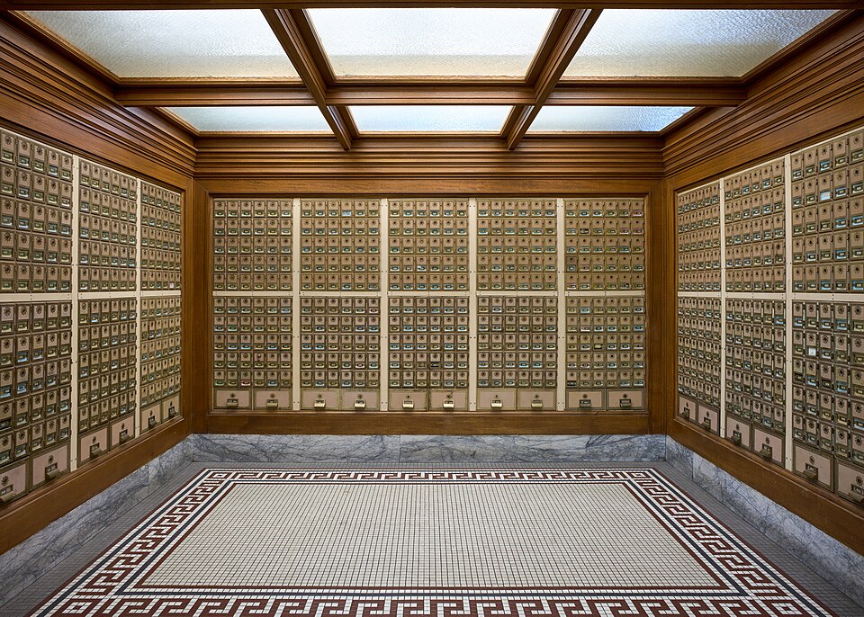 Vintage post office box alcove lined with brass mailboxes set in warm wood paneling. The floor features a white tile mosaic bordered with a black and red Greek key pattern, beneath a coffered wooden ceiling with illuminated frosted glass panels.