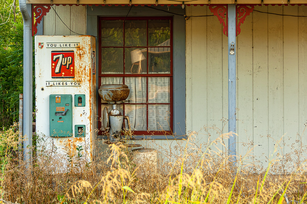 A rusty vintage 7Up vending machine and an antique cream separator stand on a porch overgrown with dry weeds beside a red-trimmed window.