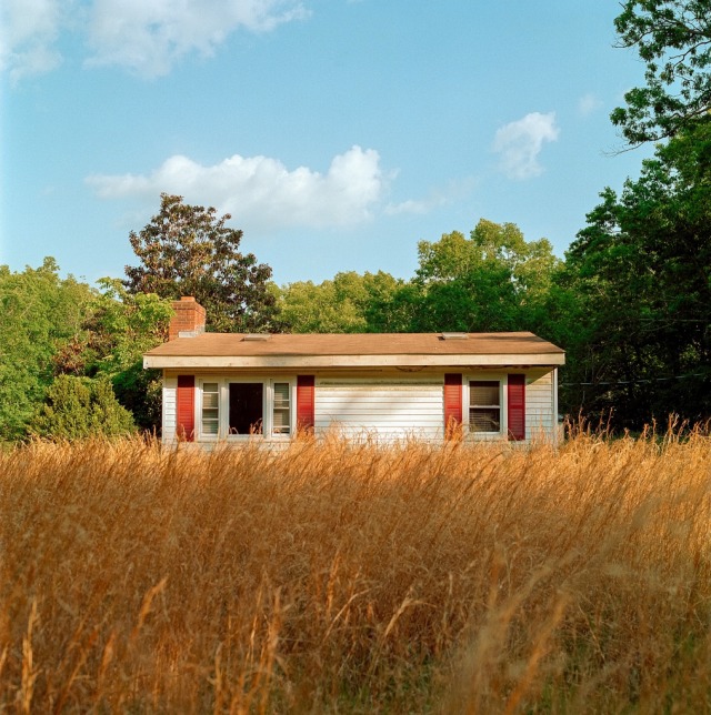 A small, single-story white house with red shutters stands behind a field of tall, golden-brown grass, backed by lush green trees under a light blue sky with scattered clouds.