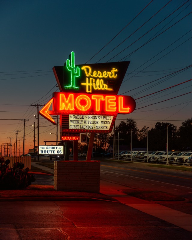 A vintage neon sign for the Desert Hills Motel glowing brightly at dusk along a roadside. The sign features a green saguaro cactus, yellow script lettering, and a large red arrow that reads "MOTEL" above an amenities reader board.